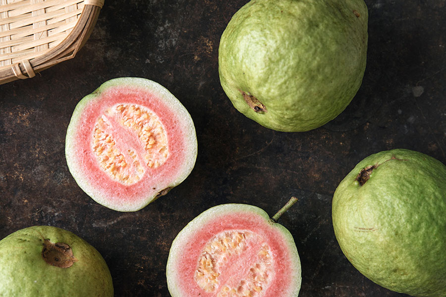 Guava Fruit - 4 guava fruits over a table - one of them is opened. Light green outside and inside its pulp is light red smooth and full of seeds.